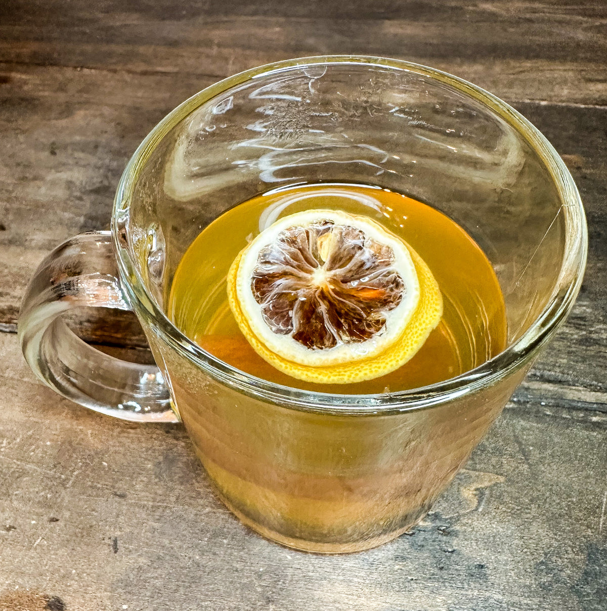 A clear glass mug filled with chamomile tea and topped with a dried lemon slice, set on a rustic wooden surface.