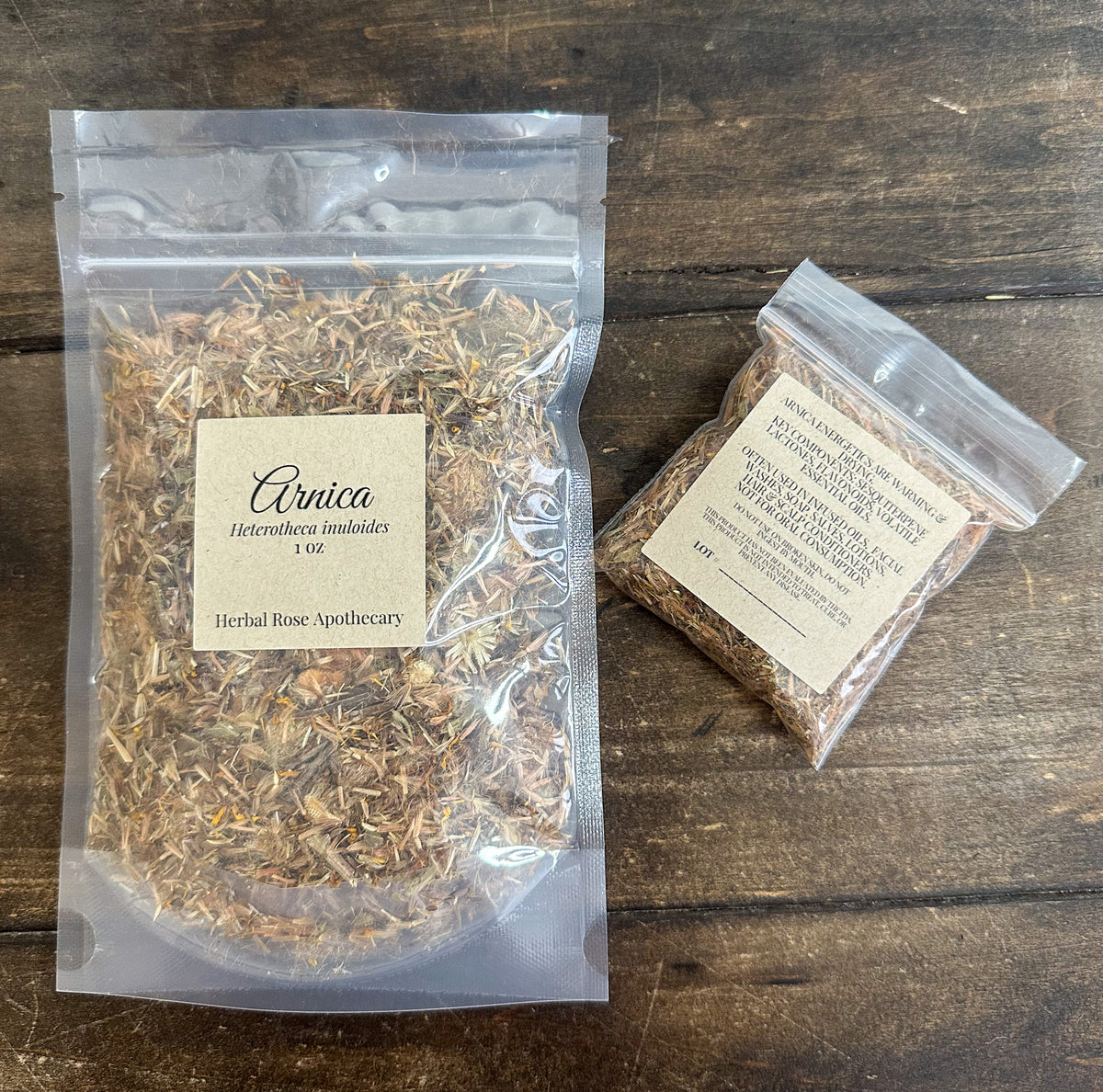Two clear resealable bags of dried botanicals on a dark wood surface. The larger bag on the left is labeled 'Arnica 1 oz' by Herbal Rose Apothecary, and the smaller bag on the right is labeled
