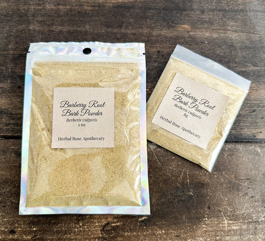 Two small bags of Barbary Reef Bark Powder by Herbal Rose Apothecary are displayed on a dark rustic wooden surface. The larger bag on the left features an iridescent silver holographic pouch with a cream-colored label, while the smaller bag on the right has a clear bag with a matching cream label.