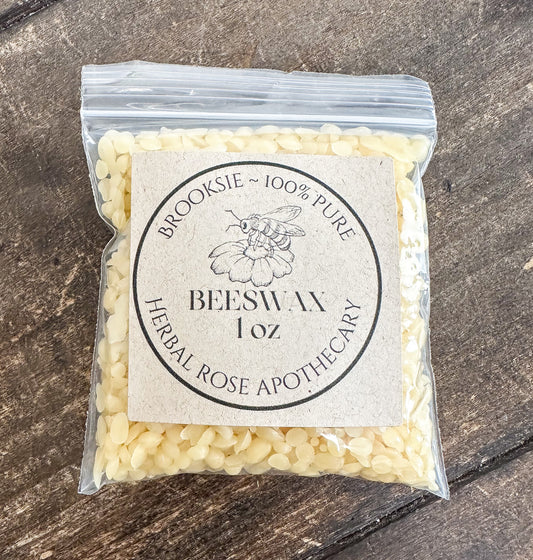 A clear plastic bag filled with pale cream beeswax pellets, featuring a round kraft paper label that reads 'Brooksie ~ 100% Pure Beeswax 1 oz Herbal Rose Apothecary,' placed on a dark rustic wooden surface.