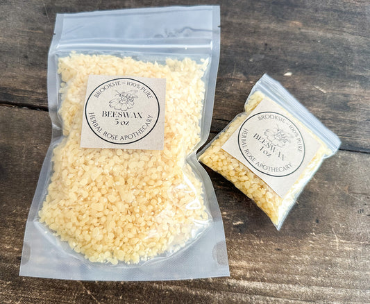 Two clear plastic bags of Beeswax flakes on a dark rustic wooden surface. The larger bag on the left contains pale yellow flakes with a square cream label reading 'Beckonside - 100% Pure BEESWAX flakes' and 'THERMAL ROSE APOTHECARY'.