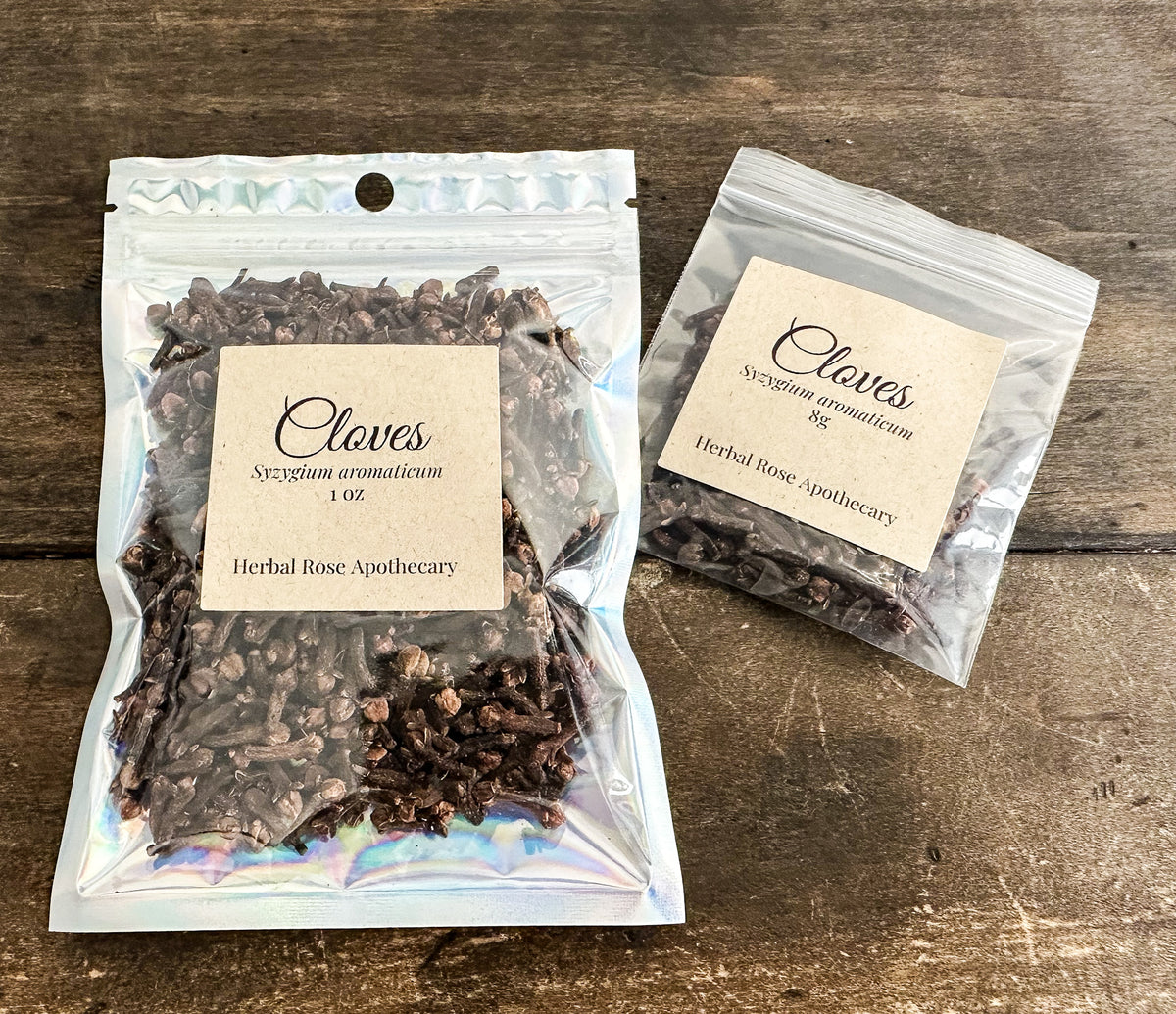 Two clear resealable pouches of cloves herbal supplement on a rustic wooden surface. The larger pouch in front is labeled 'Cloves Syzygium aromaticum 1 oz' and the smaller pouch behind is labeled 'Cloves Syzygium aromaticum 2 oz'.