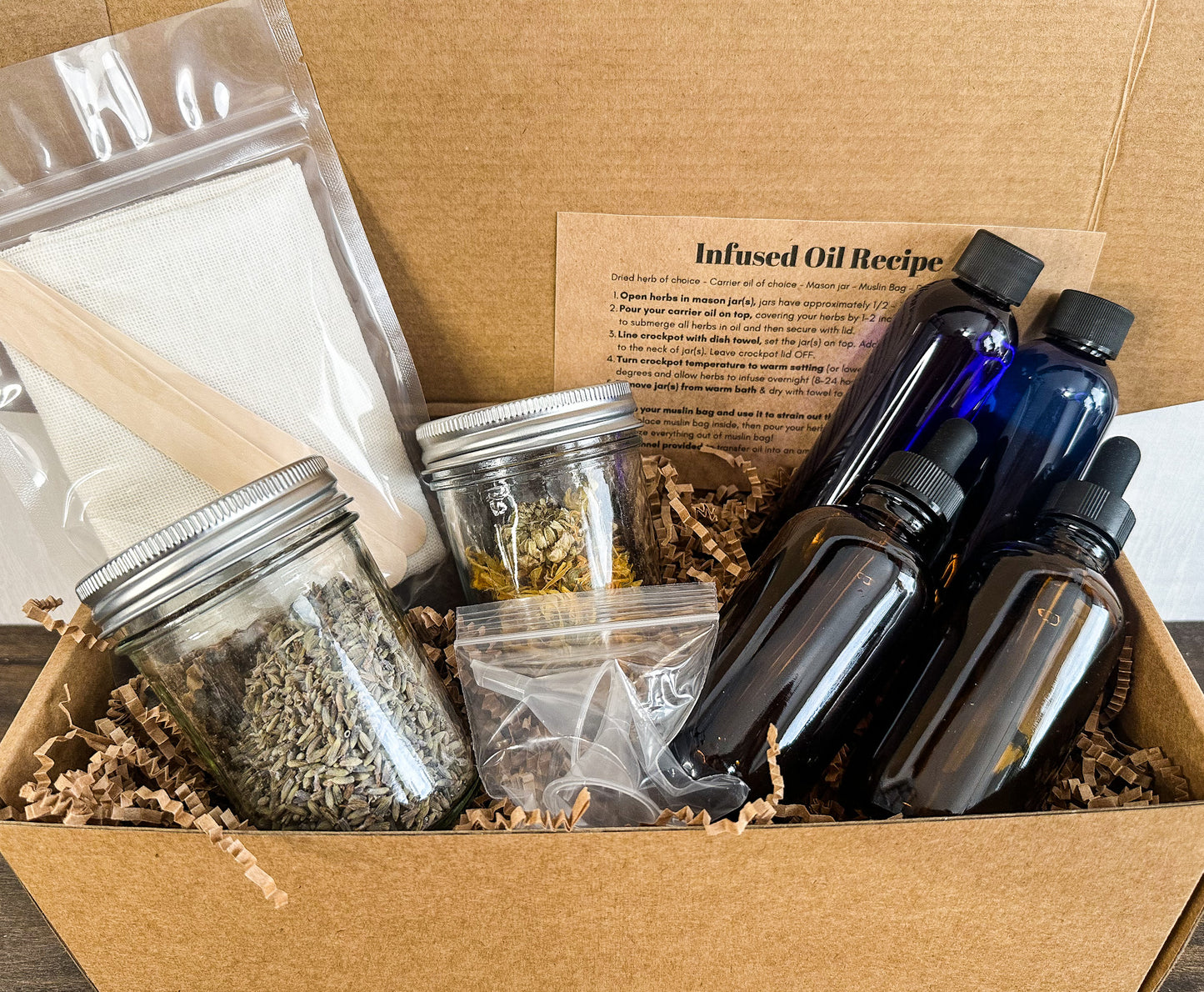 A cardboard gift box filled with a DIY infused oil kit, including dark amber glass dropper bottles, a mason jar with dried lavender, a small glass jar with dried botanicals, a clear plastic bag of white wax or soap, a wooden stir stick, and an 'Infused Oil Recipe' card.