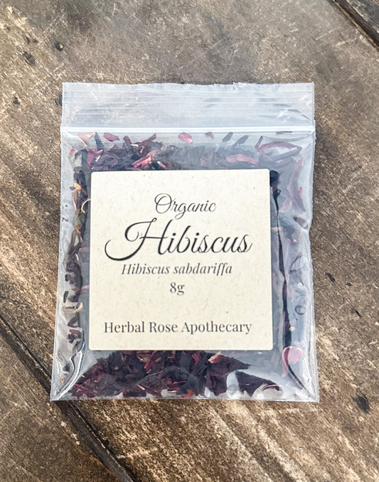 A 8g clear plastic resealable pouch of Organic Hibiscus tea by Herbal Rose Apothecary, featuring a cream-colored label, placed on a rustic wooden surface.