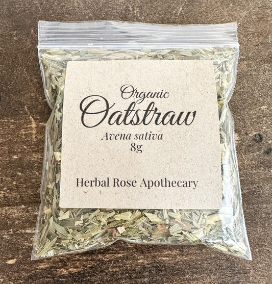 A clear plastic bag filled with dried oatstraw herbal tea, featuring a square kraft paper label that reads 'Organic Oatstraw Avena sativa 8g Herbal Rose Apothecary', set against a dark rustic wooden background.