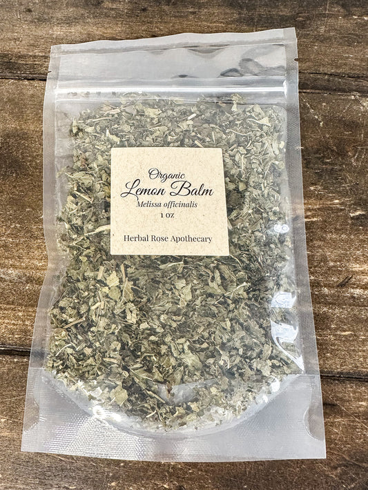 A clear stand-up pouch filled with organic dried lemon balm leaves, featuring a cream paper label that reads 'Organic Lemon Balm 1oz' and 'Herbal Rose Apothecary,' placed on a rustic wooden surface.