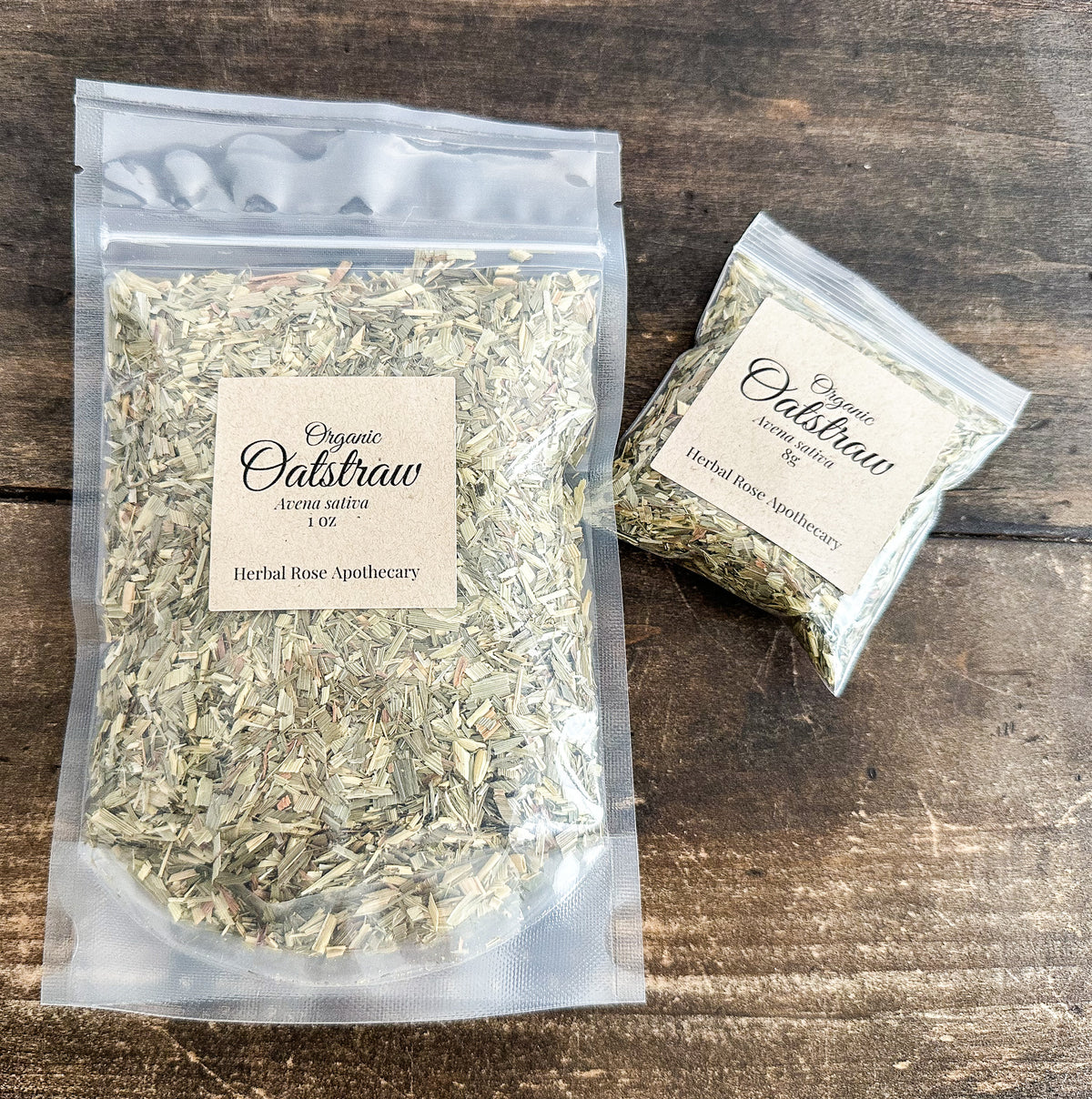 Two clear plastic bags of Organic Oakstraw Herbal Rose Apothecary tea bags placed on a dark wood surface. One larger 1 oz bag and one smaller 1/4 oz bag, both with beige paper labels.