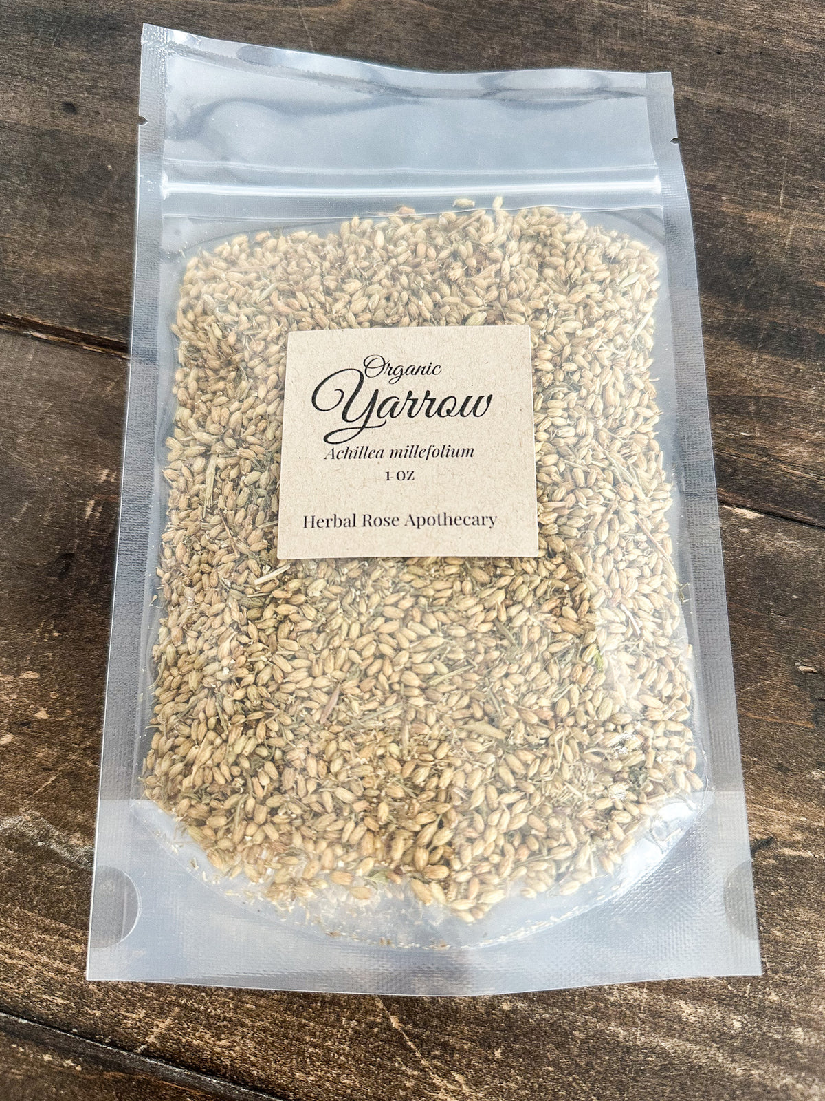 A clear stand-up pouch filled with light tan herbal grains or seeds, with a cream label reading 'Organic Yarrow' and 'Herbal Rose Apothecary,' placed on a dark rustic wooden surface.