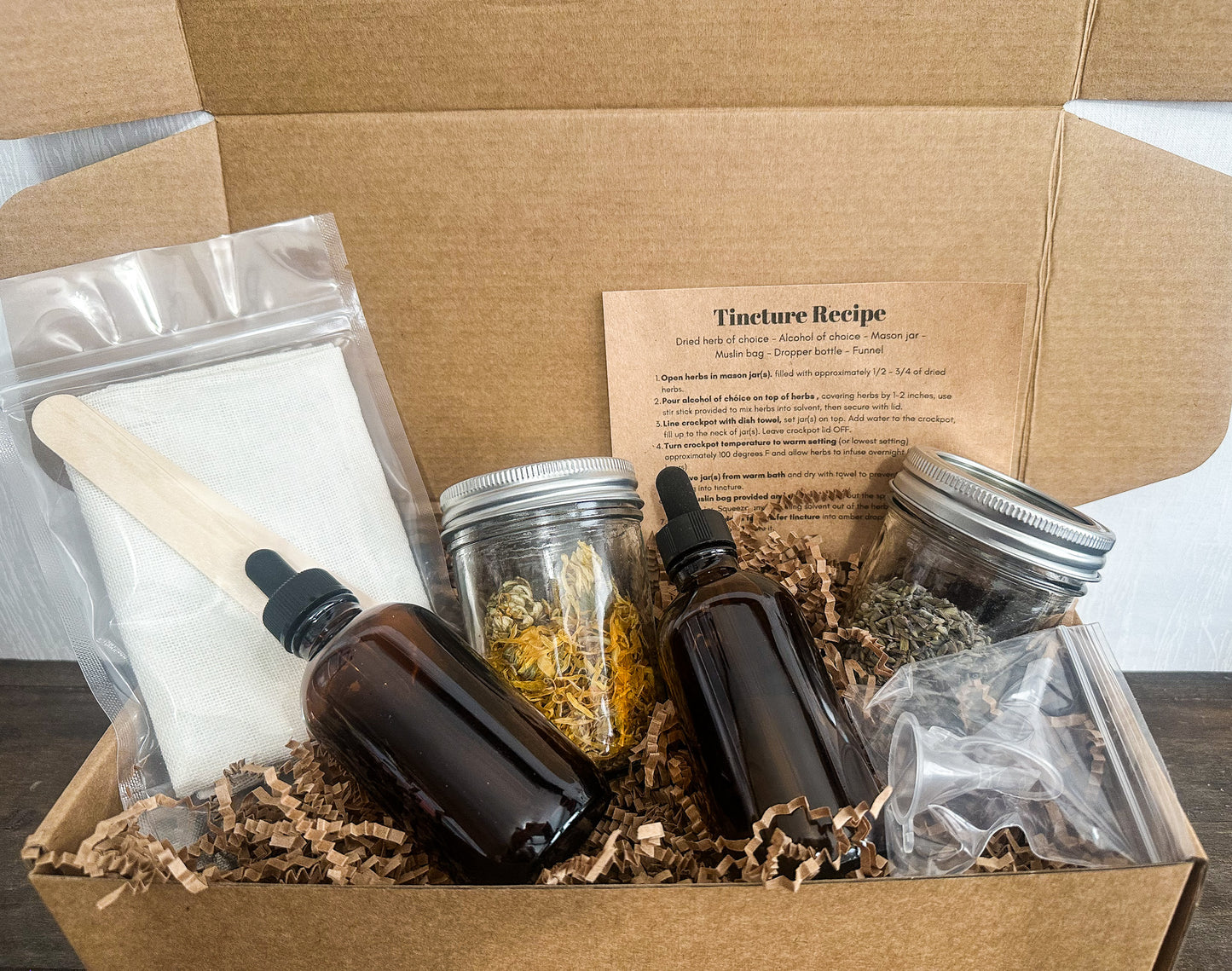 DIY tincture kit in brown kraft box showing all supplies included in box kit; cheesecloth, stir sticks, amber glass bottles, and 8oz mason jars filled half way with dried herbs, 2 funnels and instruction card