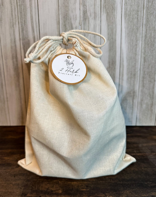 large canvas muslin bag with Kraft label hanging from standing bag, 2 mason jars, 2 amber dropper bottles, muslin bag and stir sticks in front of bag