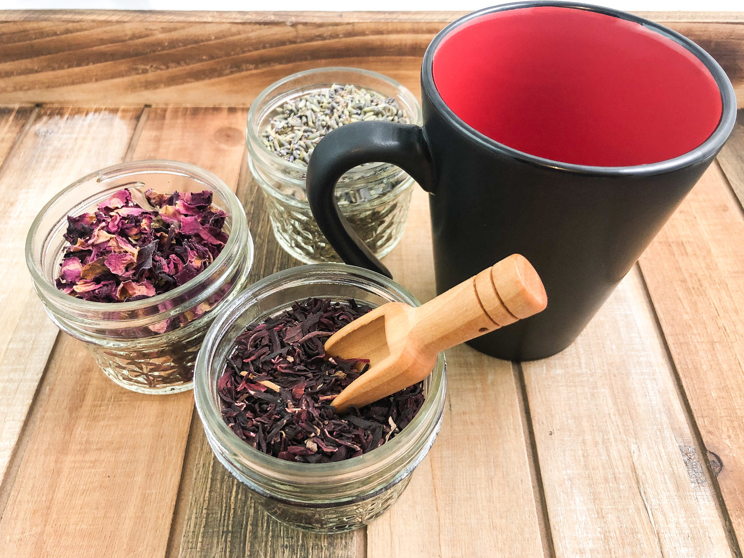 Ariel view of 3 mini glass jars filled with herbs, wooden spoon in herb jar in forefront, next to a mug with a black outside coating and red inside on a wooden background