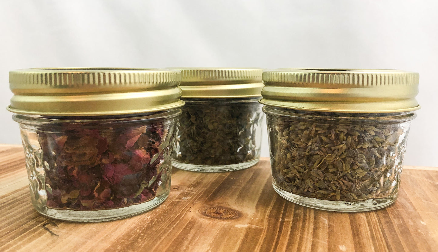 straight ahead image of mini glass clear jars filled with herbs with metal lids attached, white background on a wooden table