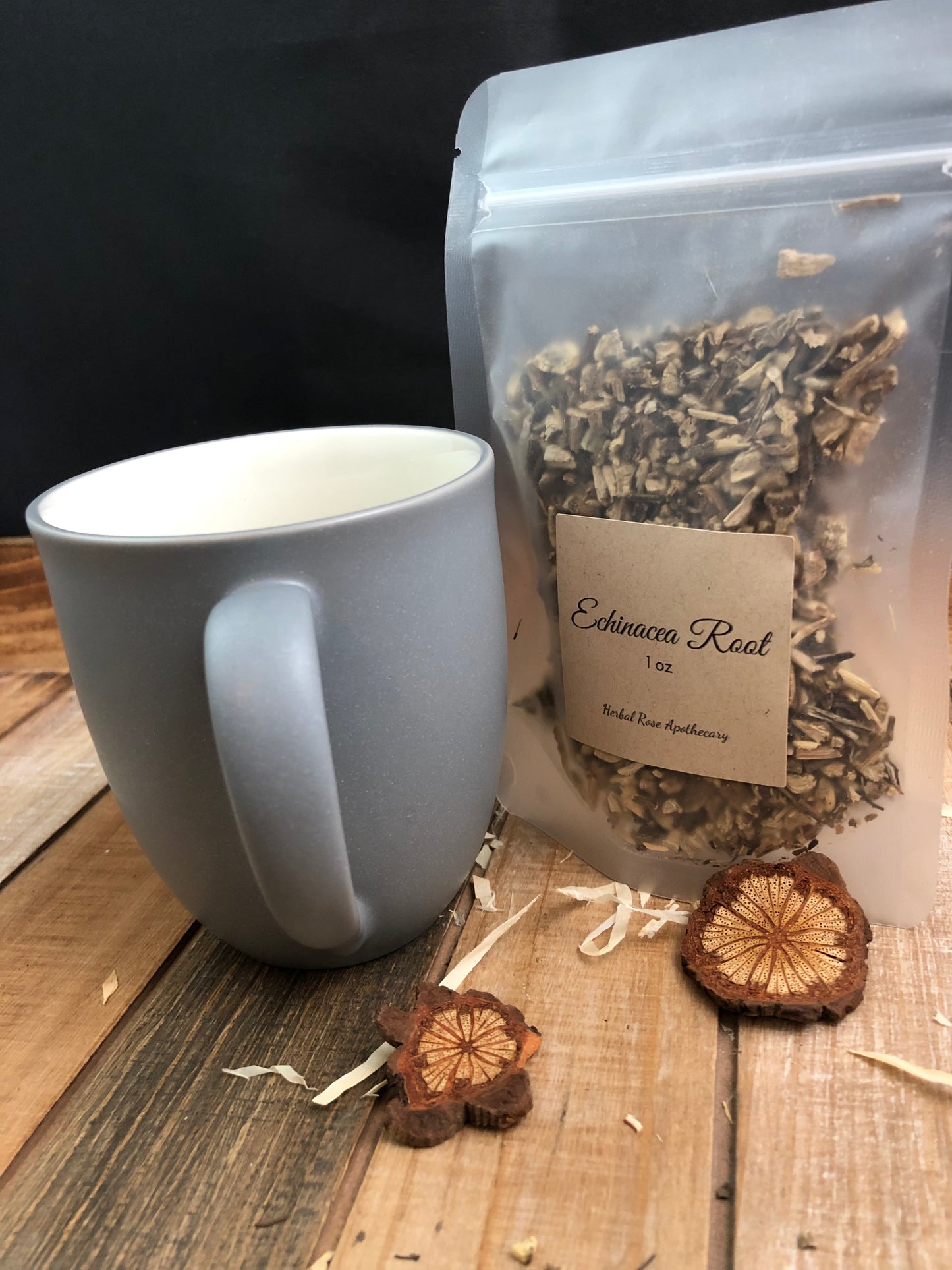 dried echinacea root in a clear plastic bag next to a grey mug and two wood chips in forefront, with white background on a wooden table