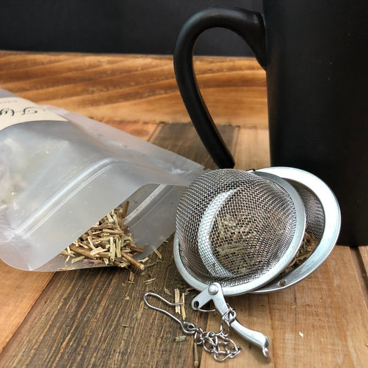 image of dried hyssop spilling out a clear bag next to a black and red coffee mug and mesh tea infuser on a wooden table with a black background