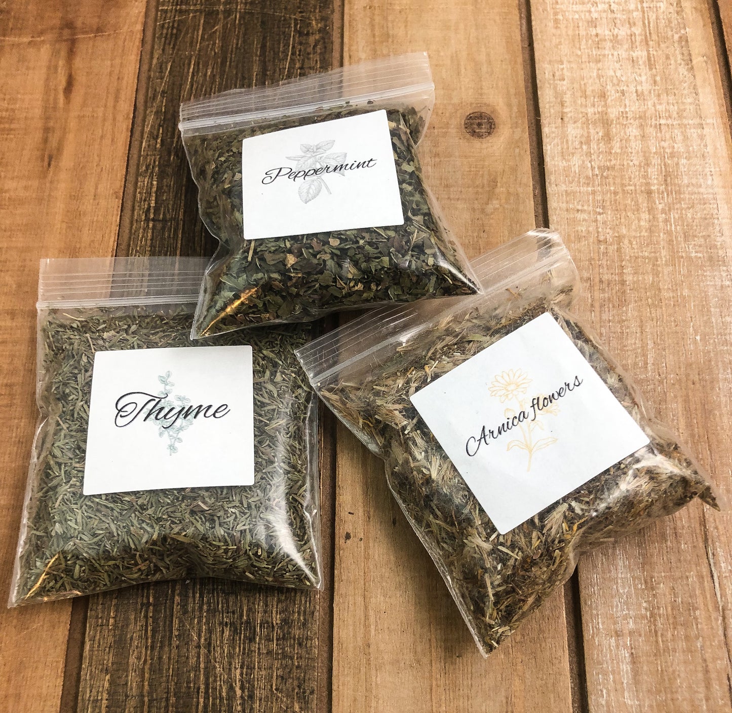 8g clear plastic bags of dried peppermint, thyme, and arnica flowers on a wooden background