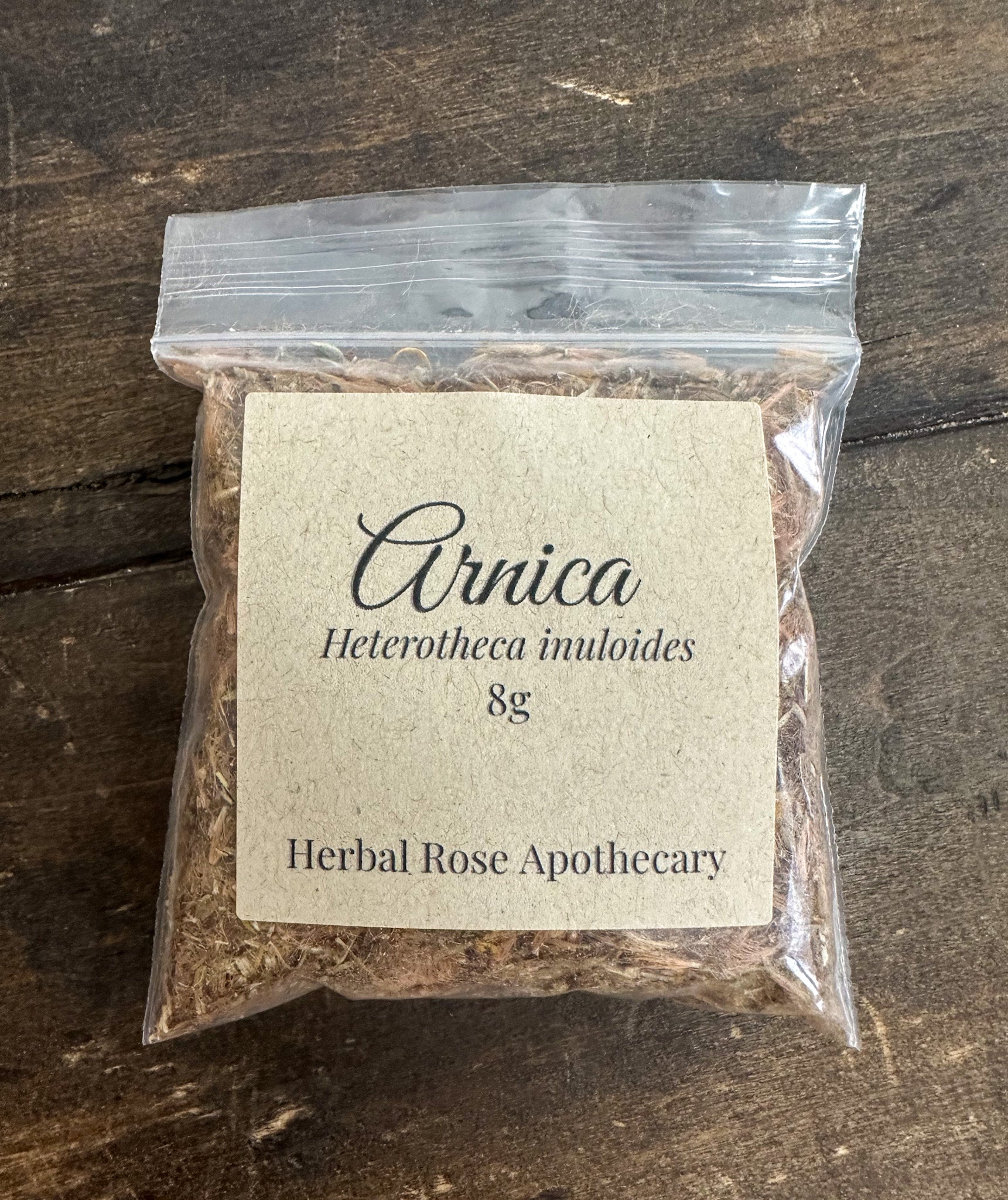Packaged herbal product labeled 'Arnica 8g' from Herbal Rose Apothecary on a wooden surface.