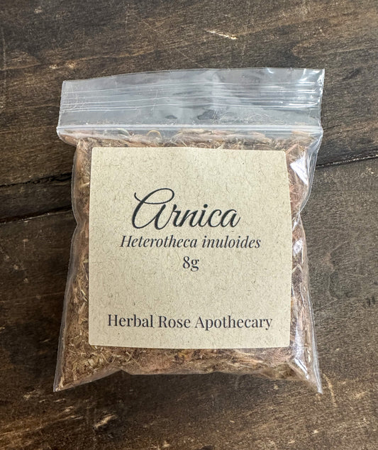 Packaged herbal product labeled 'Arnica 8g' from Herbal Rose Apothecary on a wooden surface.