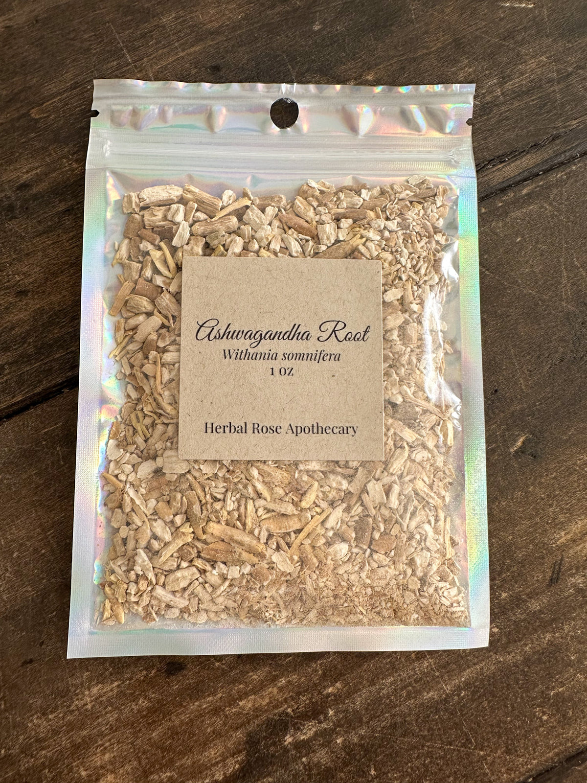 A clear holographic  pouch containing 'Ashwagandha Root 1 oz Herbal Rose Apothecary,' placed on a dark wood surface.