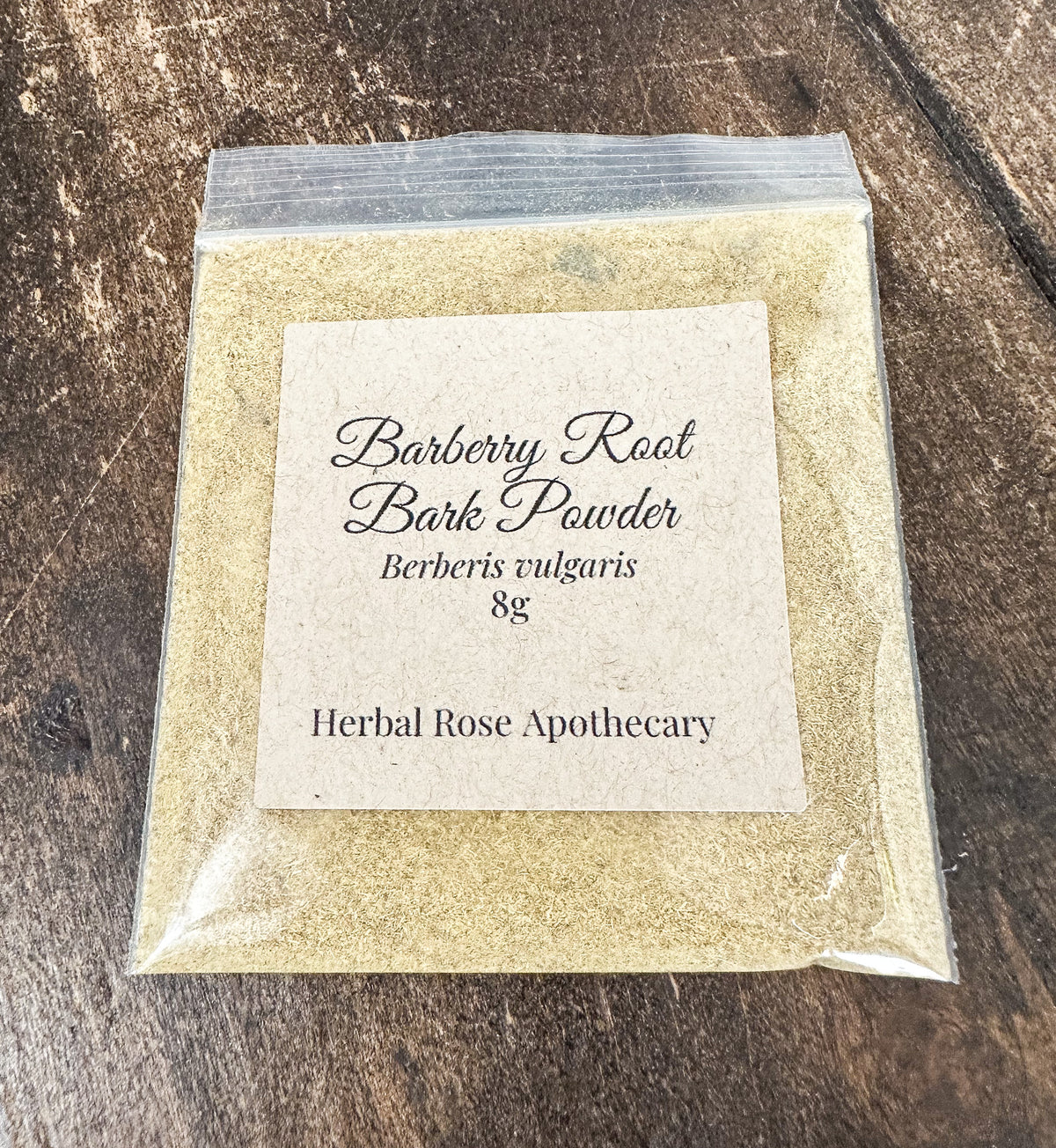 A small clear plastic bag filled with light tan barberry root bark powder sits on a dark wood surface. A cream paper label on the front reads 'Barberry Root Bark Powder', 'Berberis vulgaris', '8g', and 'Herbal Rose Apothecary'.