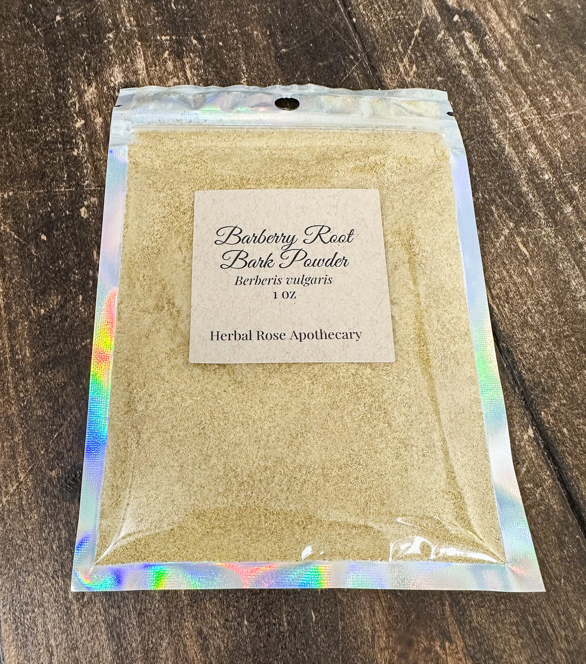 A 1 oz bag of Herbal Rose Apothecary Barberry Root Bark Powder, filled with light tan powder and sealed in a holographic silver pouch, displayed on a dark wood background.