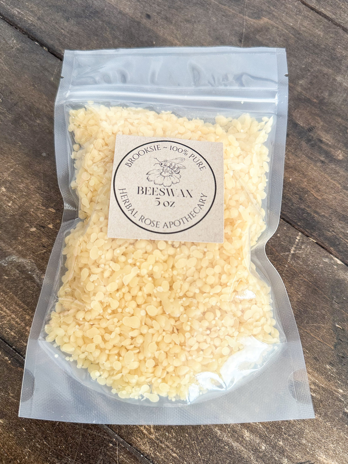 A clear plastic stand-up pouch filled with pale yellow crystalline powder, featuring a round beige label that reads 'BROOKSIE - 100% PURE BEESWAX 3 oz Herbal Rose Apothecary.' The pouch is photographed on a dark rustic wooden surface.