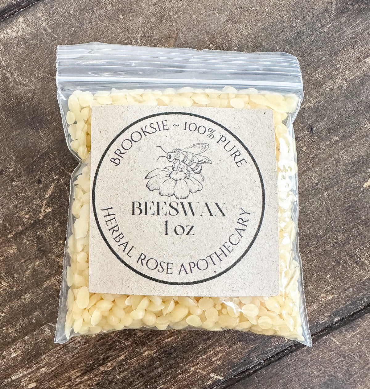 A clear plastic bag filled with pale cream beeswax pellets, featuring a round kraft paper label that reads 'Brooksie ~ 100% Pure Beeswax 1 oz Herbal Rose Apothecary,' placed on a dark rustic wooden surface.