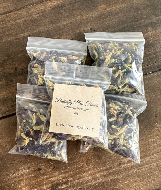 Five small clear plastic zip-top bags filled with dried butterfly pea flowers arranged on a rustic wooden surface. One bag is centered and labeled with a kraft paper tag that reads 'Butterfly Pea Flower Cichorium tenues 8g Herbal Rose Apothecary'.