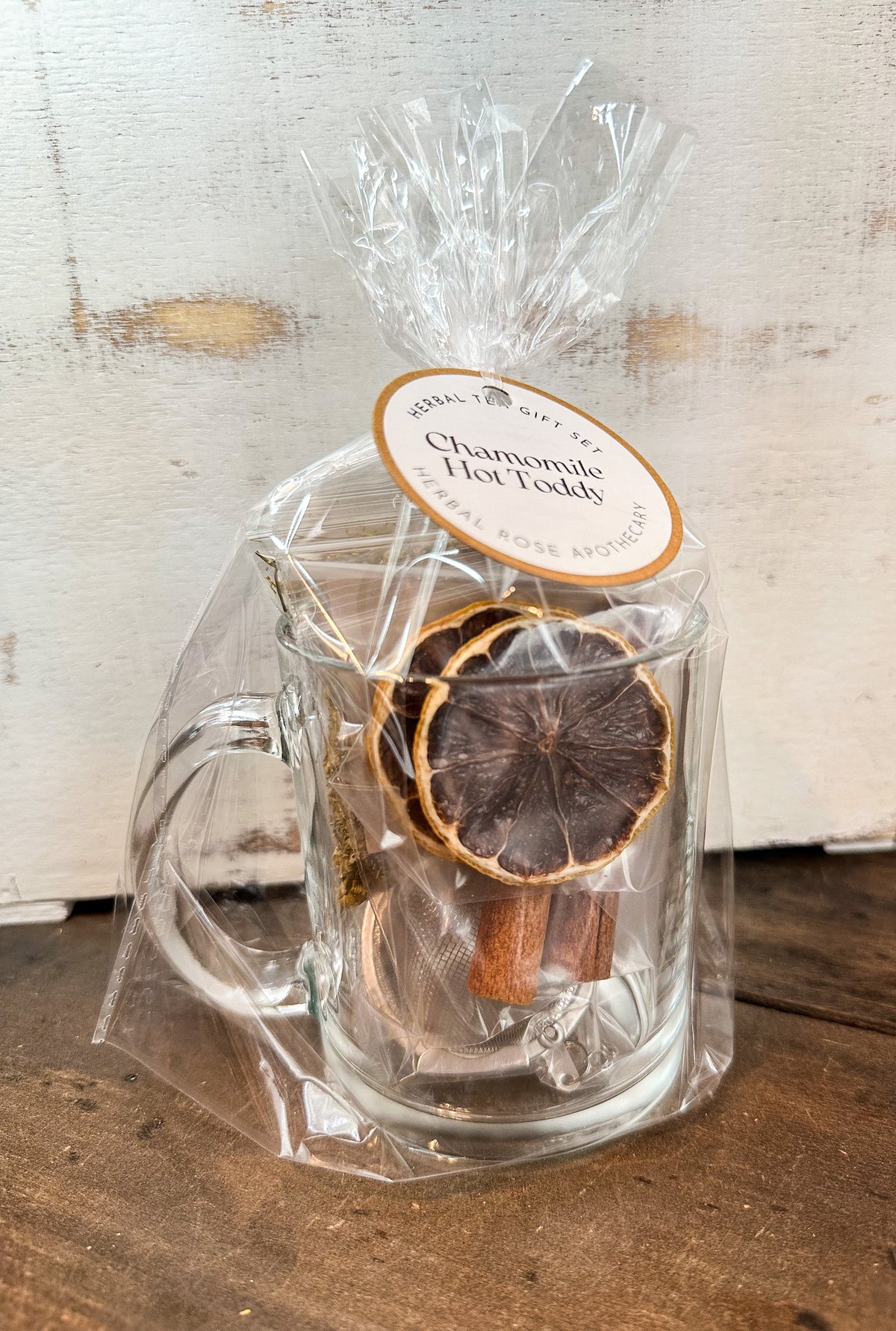 Clear glass mug with dried herbs and cinnamon sticks, wrapped in cellophane with a label on a wooden surface.