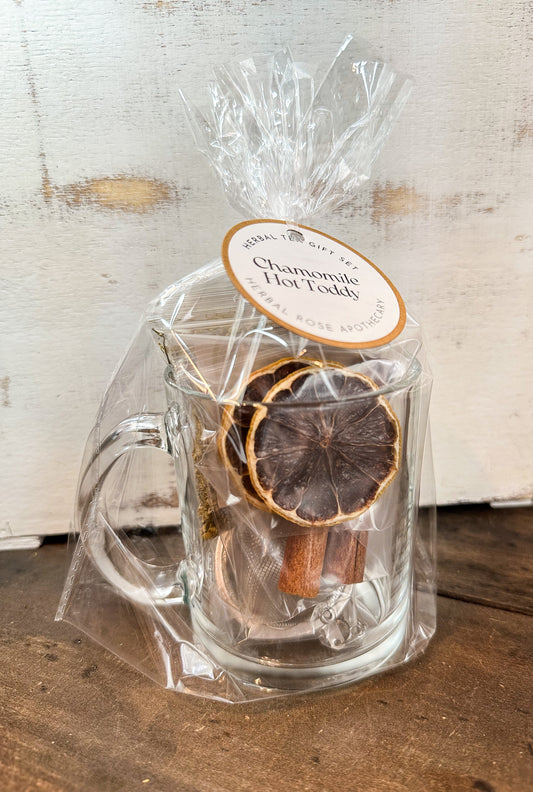Clear glass mug with dried herbs and cinnamon sticks, wrapped in cellophane with a label on a wooden surface.