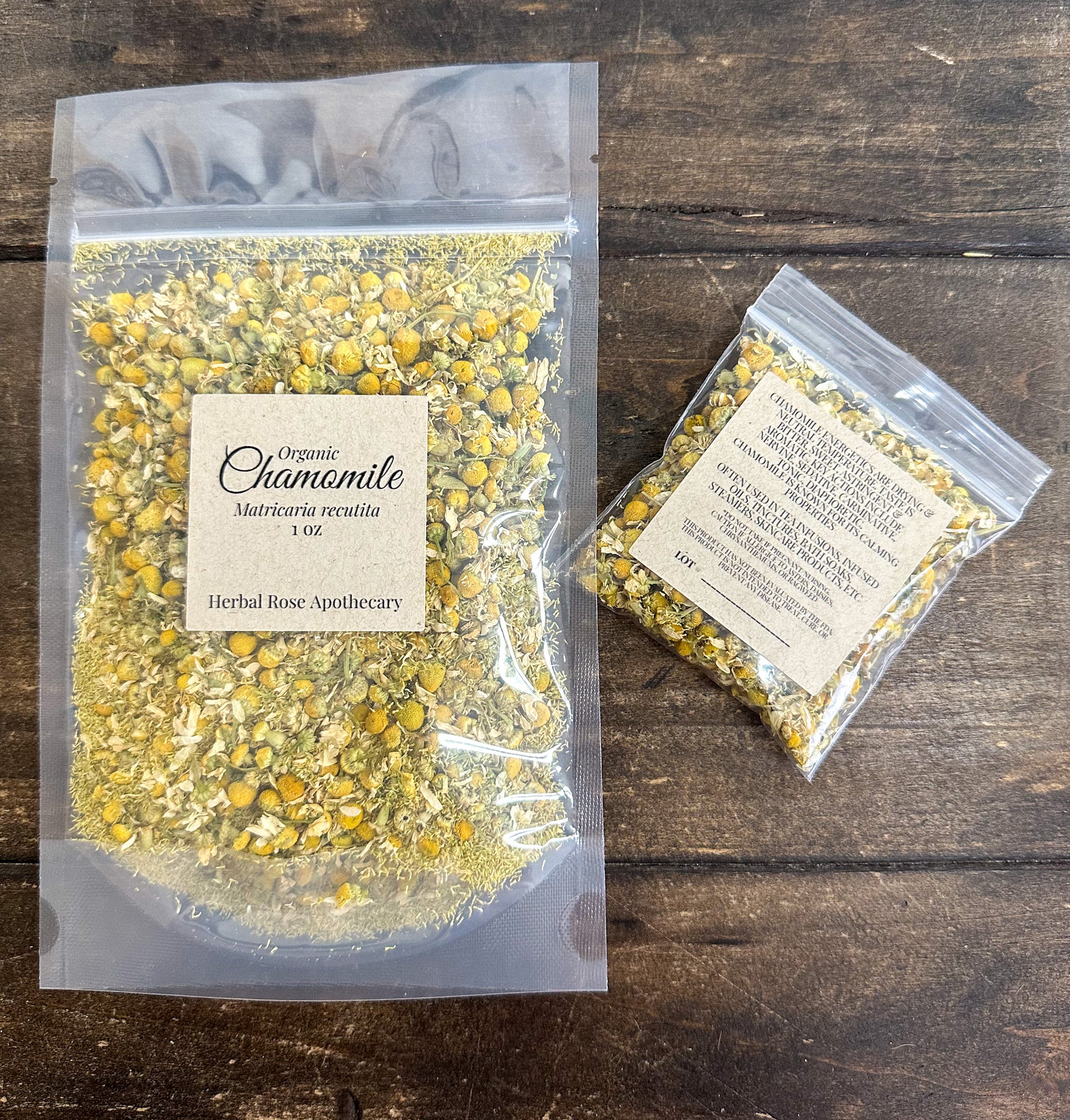 Two clear plastic resealable bags of Herbal Rose Apothecary Organic dried Chamomile, one 1oz bag and one 8g bag, displayed on a dark wood background.