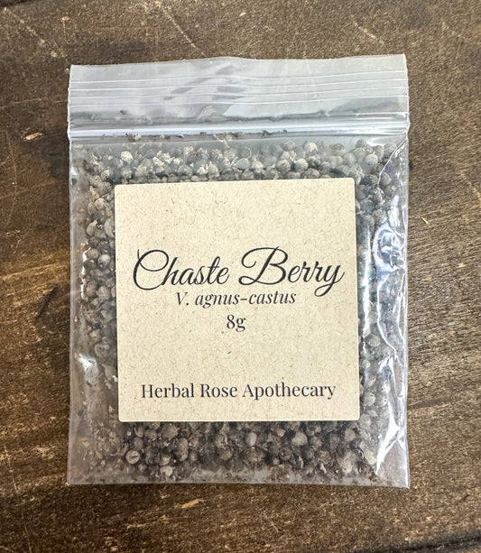 A clear plastic zip-top bag filled with chaste berries, featuring a beige paper label that reads 'Chaste Berry V. agnus-castus 8g' and 'Herbal Rose Apothecary,' placed on a dark wood surface.