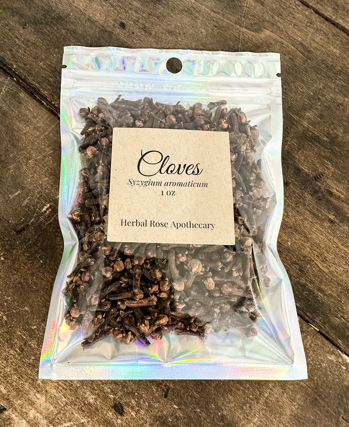 A clear holographic stand-up pouch filled with whole cloves, labeled 'Cloves Syzygium aromaticum 1 oz Herbal Rose Apothecary,' placed on a rustic wooden surface.