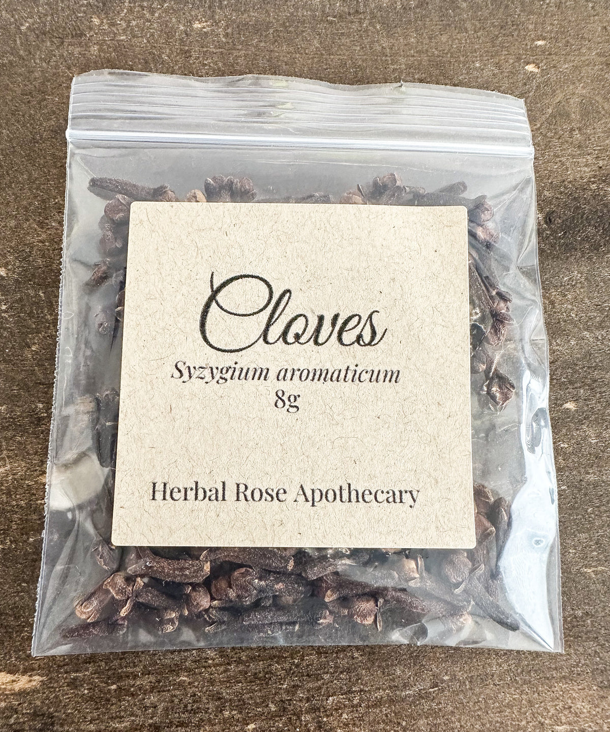 A clear resealable plastic bag containing dried cloves, with a centered kraft paper label reading 'Cloves Syzygium aromaticum 8g Herbal Rose Apothecary' on a brown textured surface.