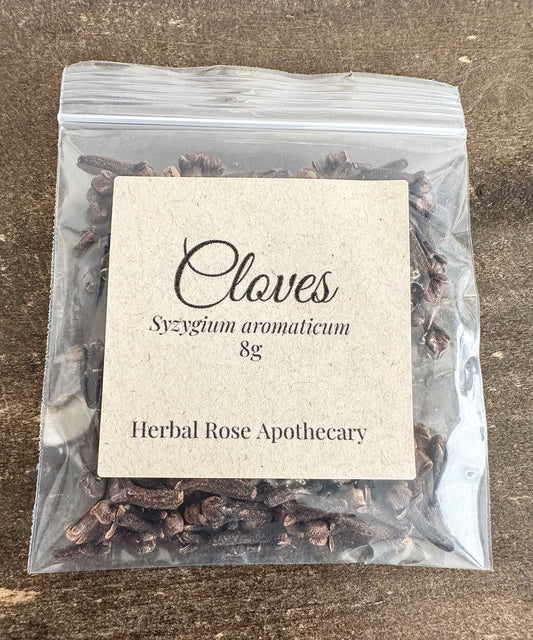 A clear resealable plastic bag containing dried cloves, with a centered kraft paper label reading 'Cloves Syzygium aromaticum 8g Herbal Rose Apothecary' on a brown textured surface.