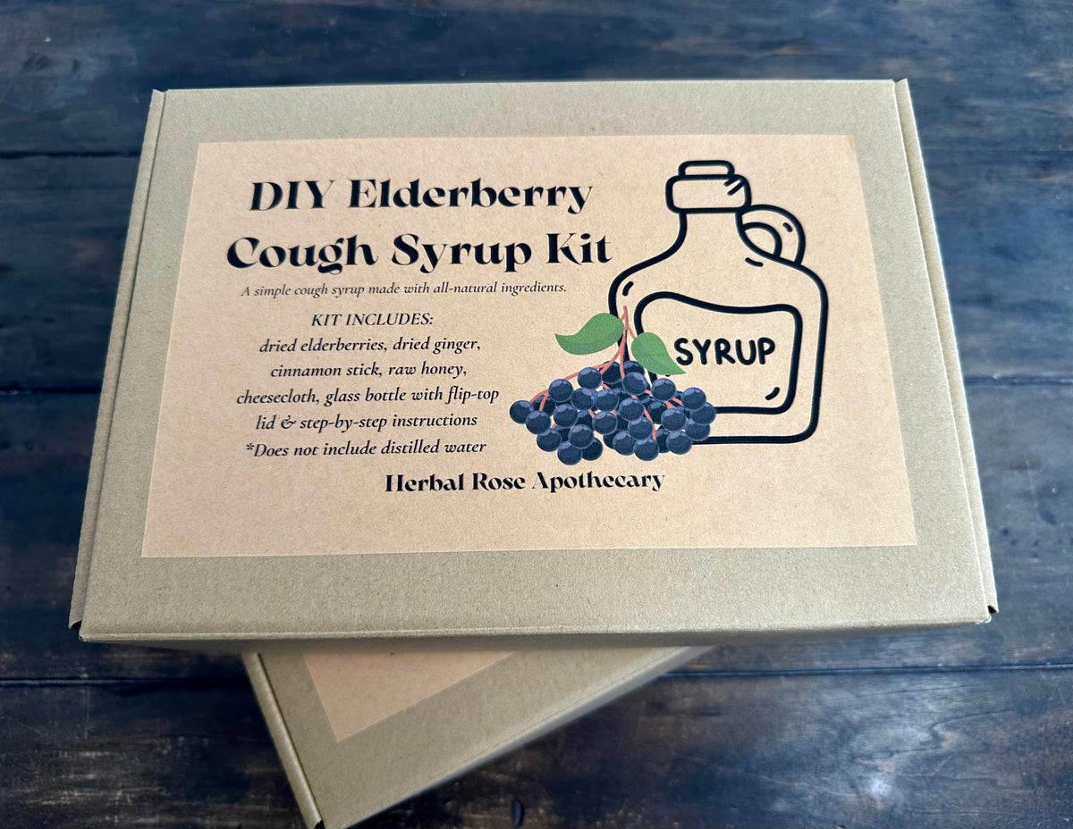 A DIY Elderberry Cough Syrup Kit box by Herbal Rose Apothecary, shown on a dark wooden surface. The box is kraft paper with a beige label featuring a black line drawing of a syrup bottle and a cluster of dark purple elderberries.