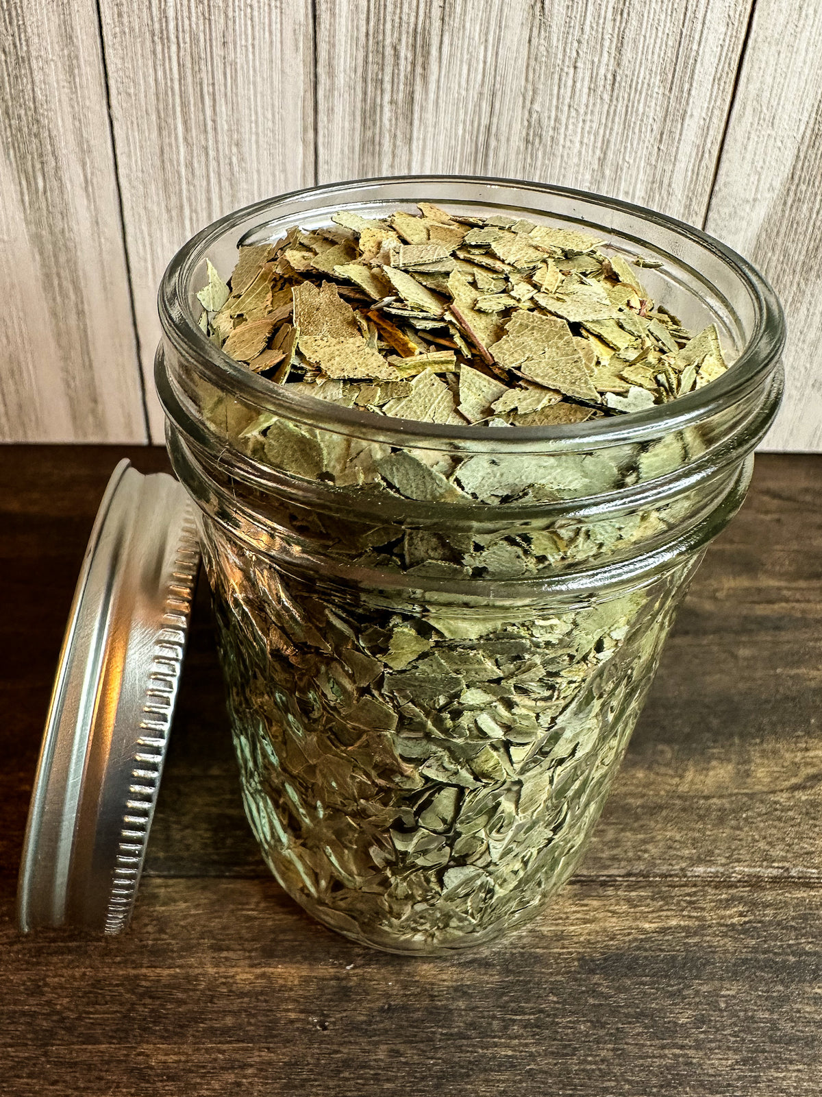 A clear glass mason jar filled with dried eucalyptus leaves, shown on a dark wooden surface with a silver metal lid resting beside it. The background features a light wood plank wall.
