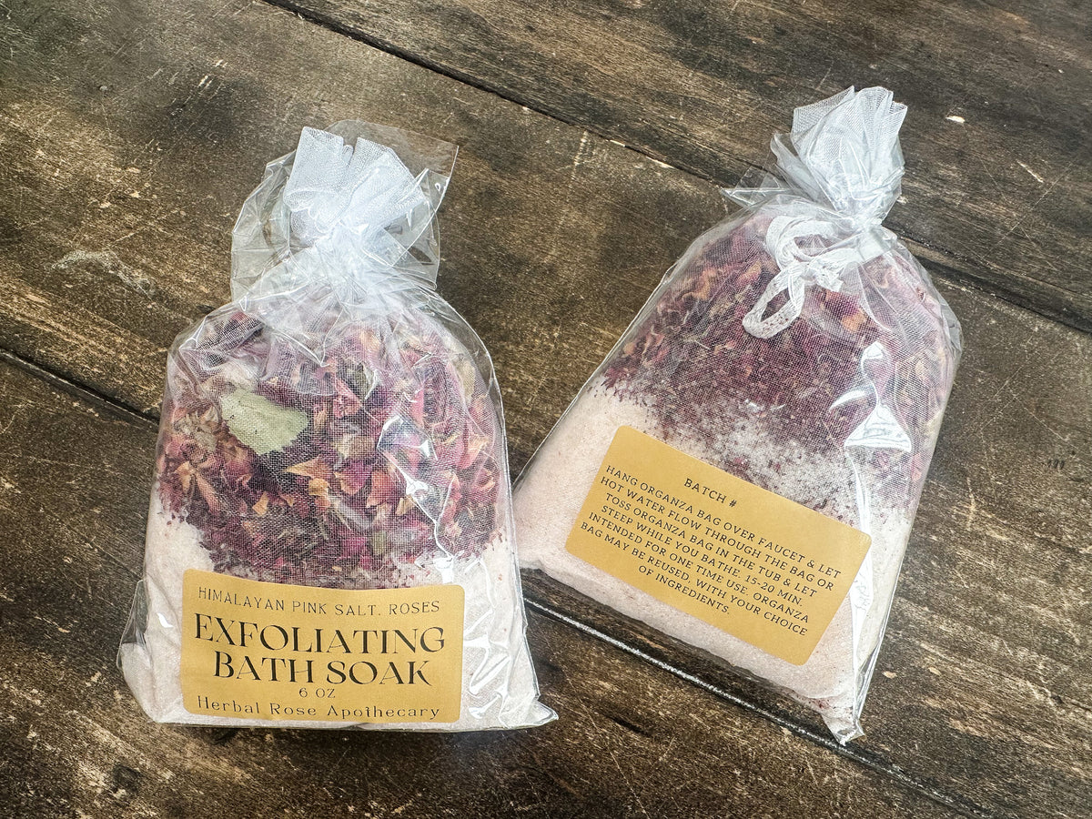 two exfoliating bath soaks packaged in a clear bag and labeled lying on a wooden surface from herbal rose apothecary
