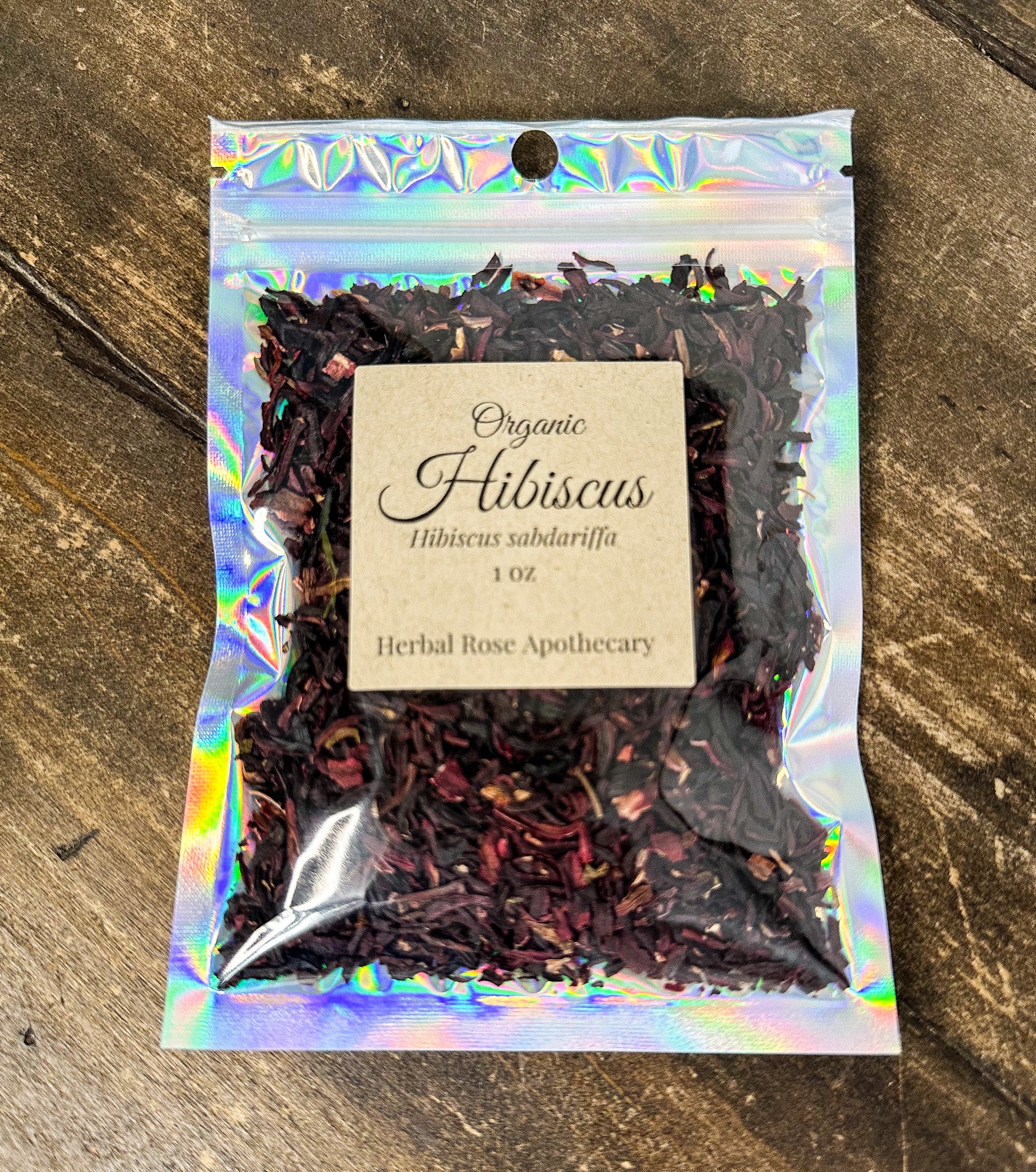A 1 oz bag of Organic Hibiscus tea from Herbal Rose Apothecary, featuring dark red hibiscus petals in an iridescent holographic stand-up pouch with a beige paper label, set on a rustic wooden background.