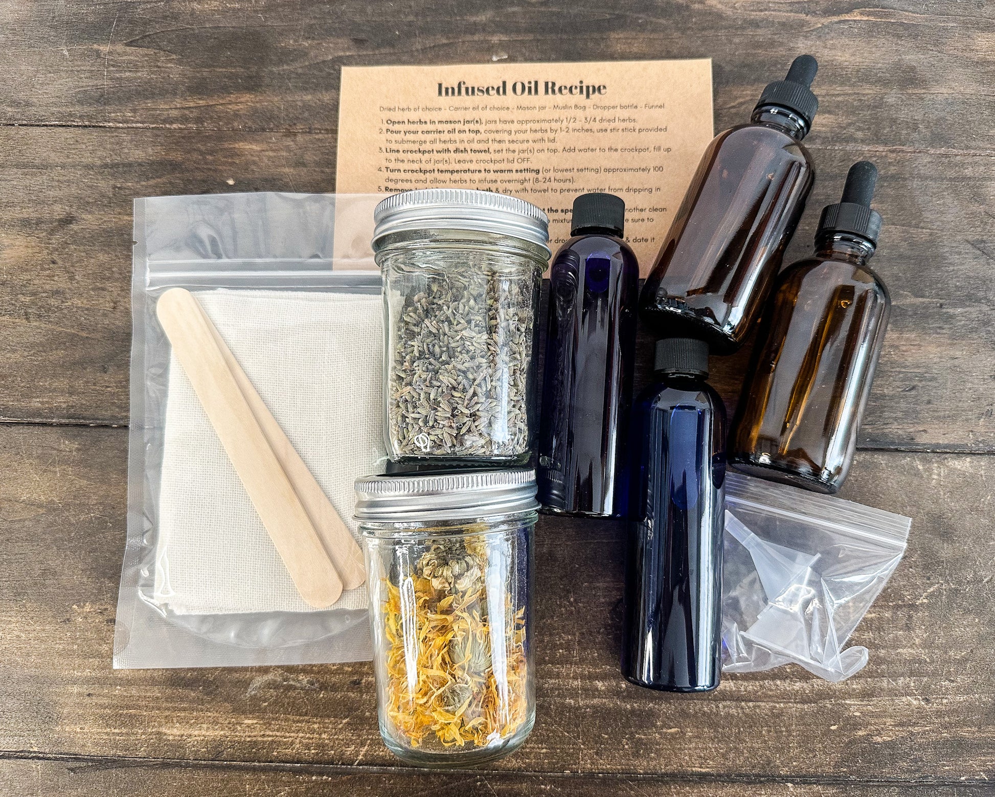 DIY infused oil box kit with supplies laid out on wooden background; infused oil recipe card, stir sticks, cheesecloths, 2 herb jars, 2 almond oil bottles, 2 amber glass bottles, and 2 funnels