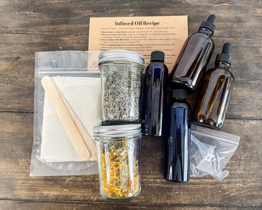 DIY infused oil box kit with supplies laid out on wooden background; infused oil recipe card, stir sticks, cheesecloths, 2 herb jars, 2 almond oil bottles, 2 amber glass bottles, and 2 funnels
