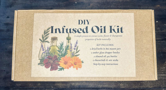 DIY Infused Oil Kit in a square kraft paper box with a cream label featuring botanical illustrations and product text, photographed from above on a dark wooden surface.