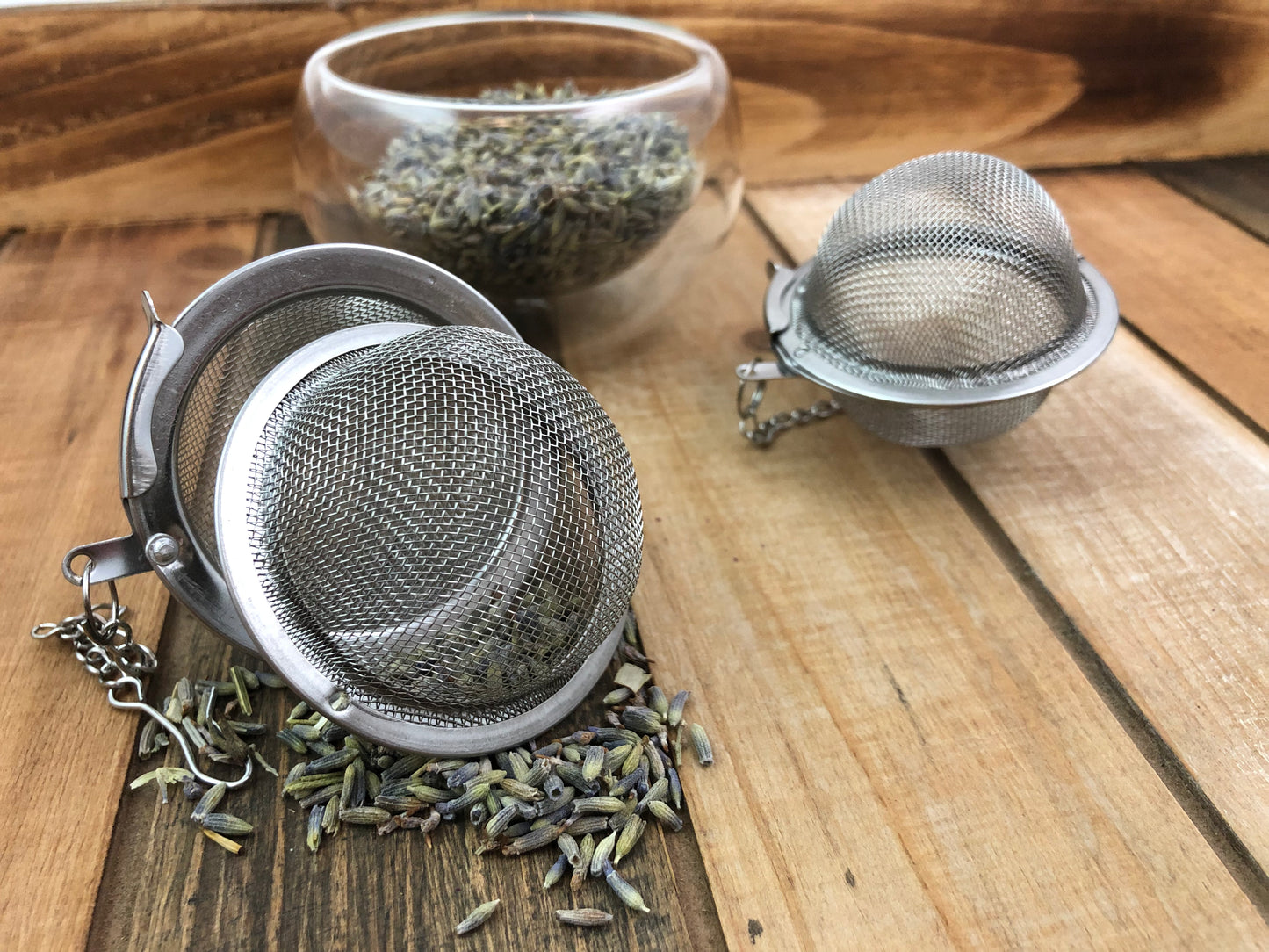 dried lavender buds spilling out of a tea ball infuser, dried lavender buds in a glass bowl and an additional tea ball infuser in the background all on a wooden surface