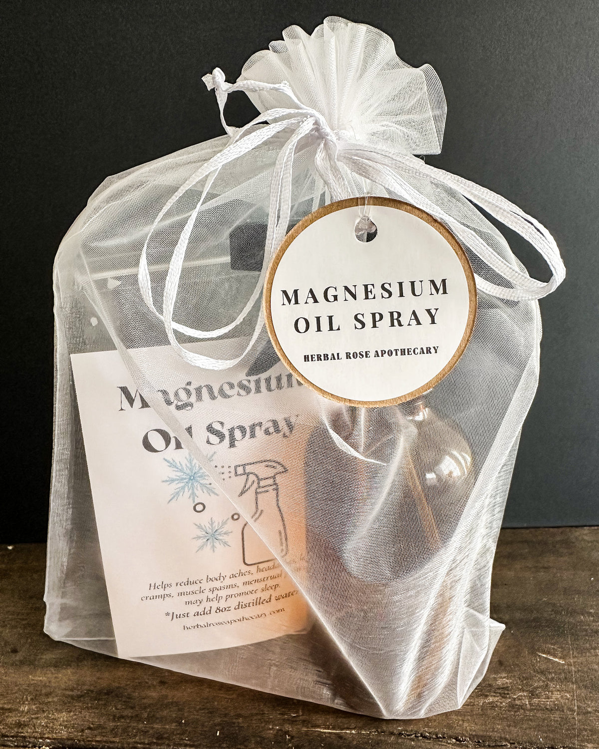 A clear mesh drawstring gift bag containing a DIY kit to make a bottle of Herbal Rose Apothecary Magnesium Oil Spray, set on a rustic wooden surface against a dark background.