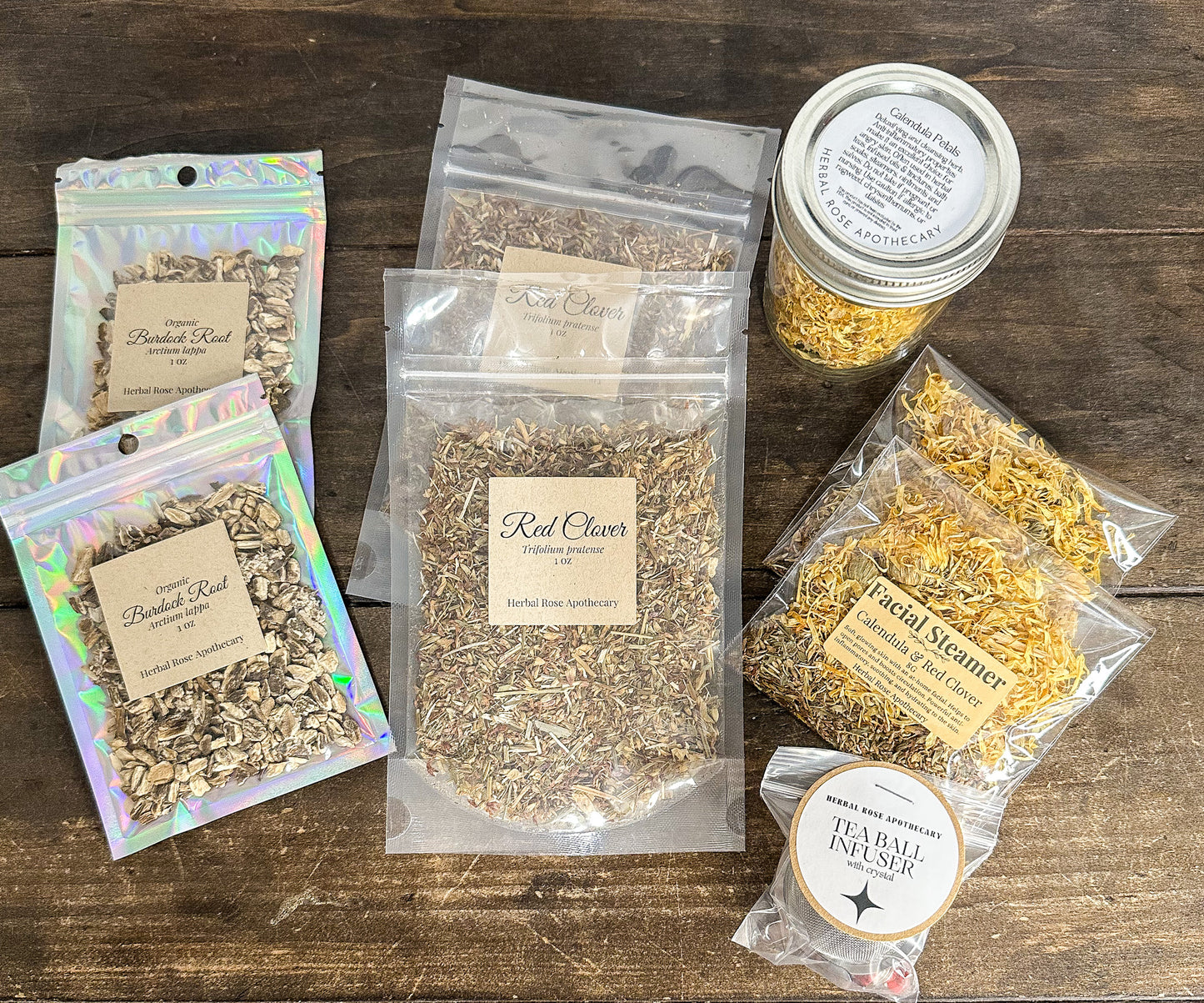 2 red clover 1oz bags, 2 burdock root 1oz bags, 1 calendula jar and 2 facial steamers with calendula & red clover, and 1 tea ball infuser on a wooden table