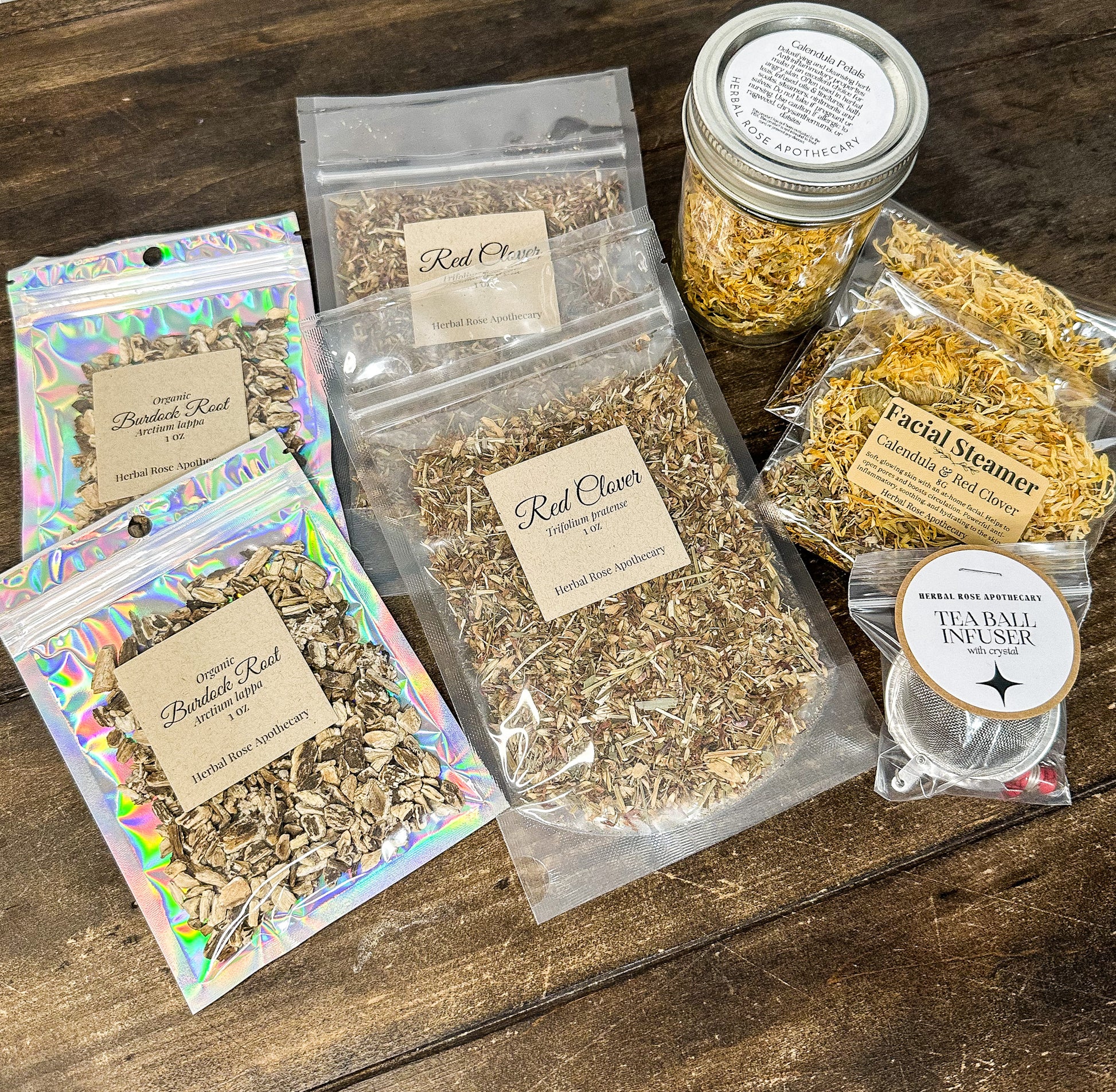 2 red clover 1oz bags, 2 burdock root 1oz bags, 1 calendula jar and 2 facial steamers with calendula & red clover, and 1 tea ball infuser on a wooden table