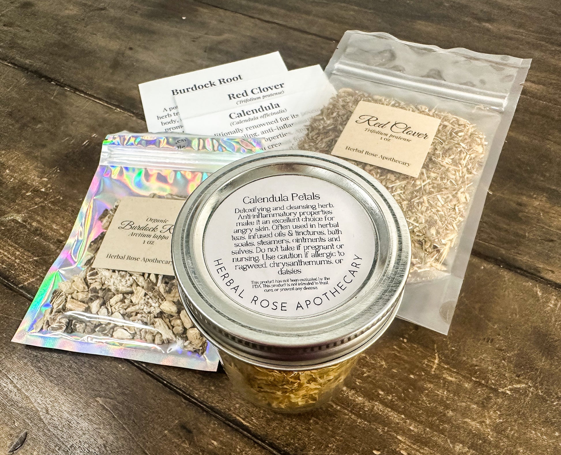 jar of calendula flowers, 1oz bag of red clover, 1 oz bag of burdock root with monograph cards for each herb lying on a wooden table