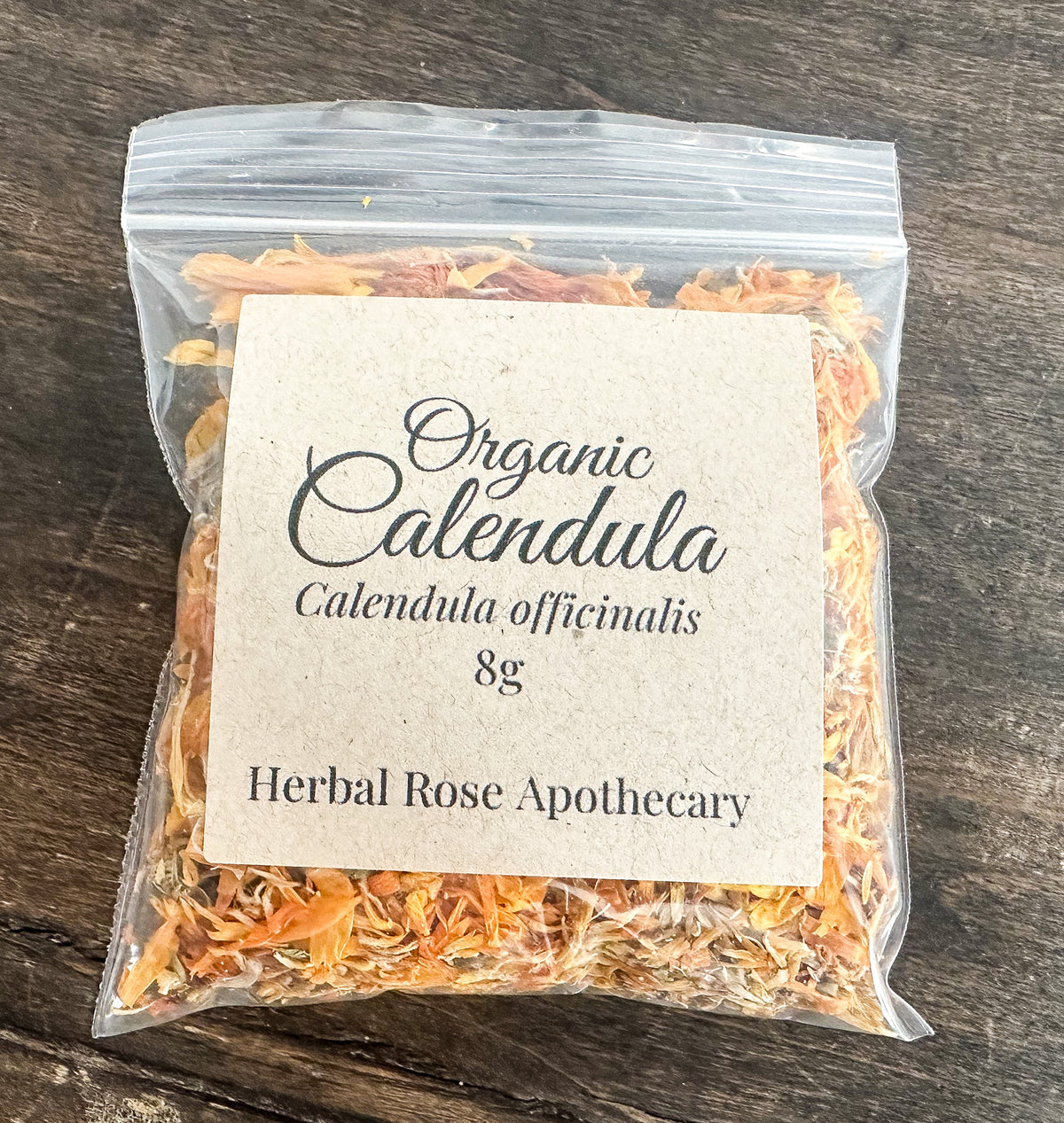 A clear plastic resealable bag filled with dried orange calendula flowers, with a large cream paper label reading 'Organic Calendula Calendula officinalis 8g Herbal Rose Apothecary' on a dark wooden surface.