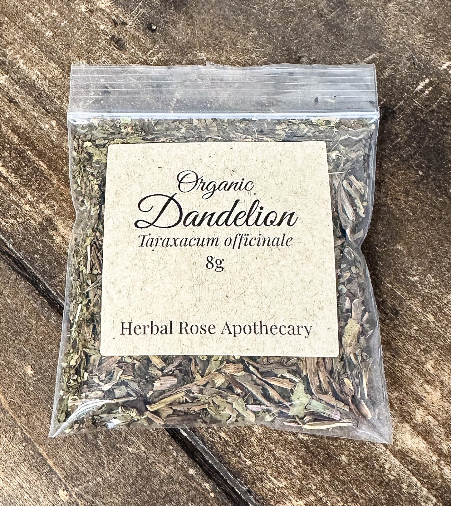 A clear plastic resealable bag filled with dried organic dandelion, featuring a cream paper label that reads 'Organic Dandelion Taraxacum officinale 8g' and 'Herbal Rose Apothecary,' placed on a rustic dark wood surface.