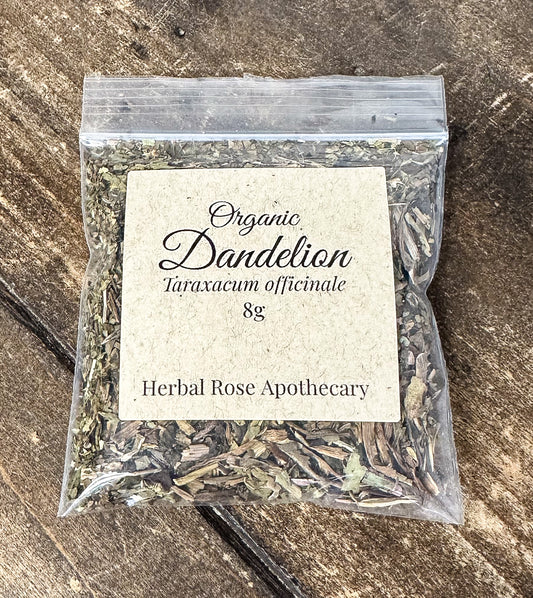 A clear plastic resealable bag filled with dried organic dandelion, featuring a cream paper label that reads 'Organic Dandelion Taraxacum officinale 8g' and 'Herbal Rose Apothecary,' placed on a rustic dark wood surface.