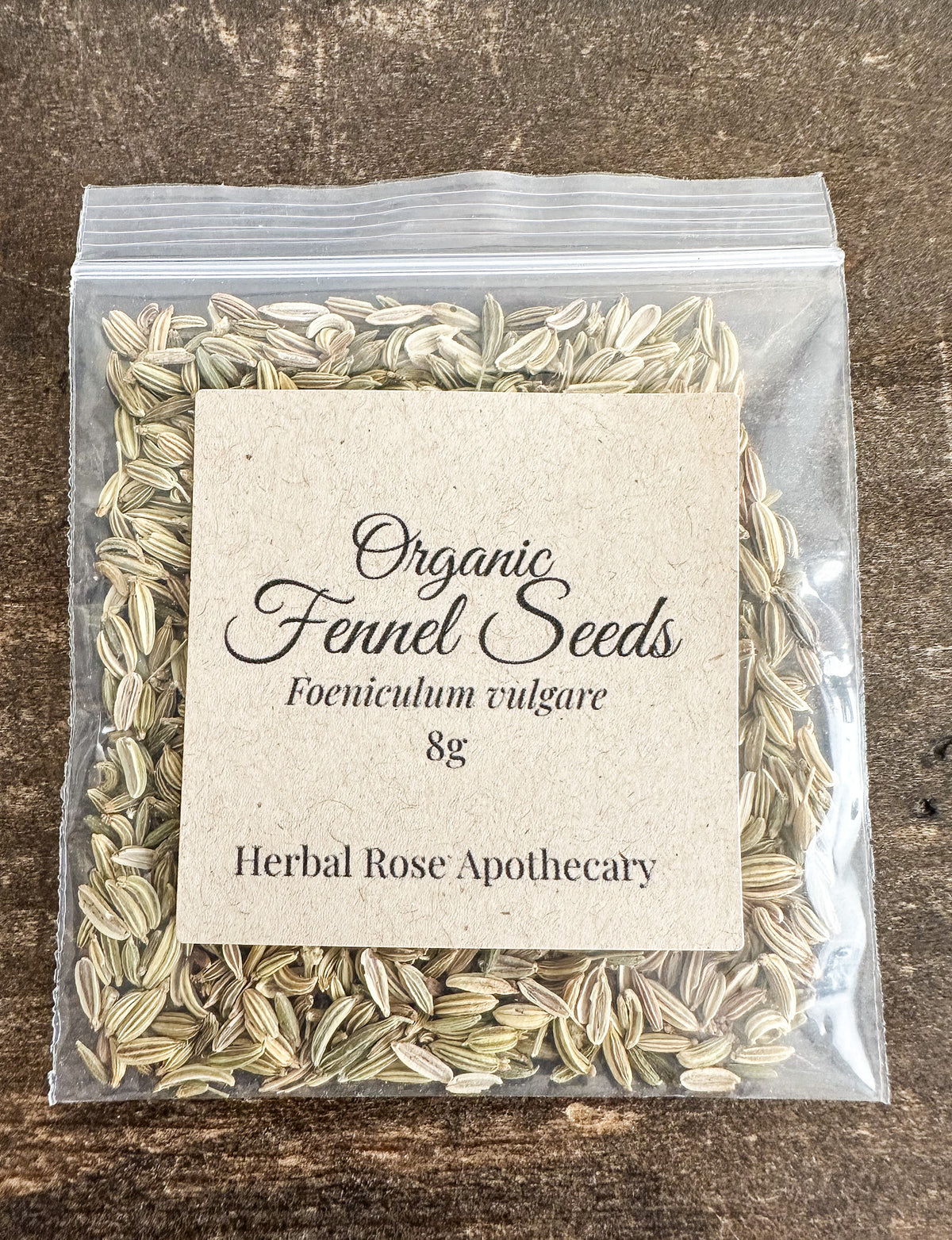 A clear plastic resealable bag filled with dried organic fennel seeds, with a centered kraft paper label reading 'Organic Fennel Seeds Foeniculum vulgare 8g Herbal Rose Apothecary,' placed on a dark wood surface.